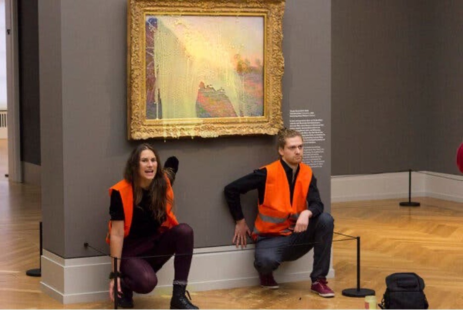 Two people in orange vests glued their hands to a wall underneath a painting.