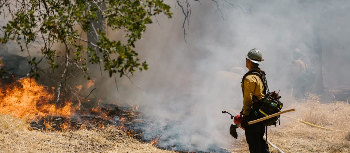 Firefighter standing on the frontlines of a wildfire with smoke blowing towards him.
