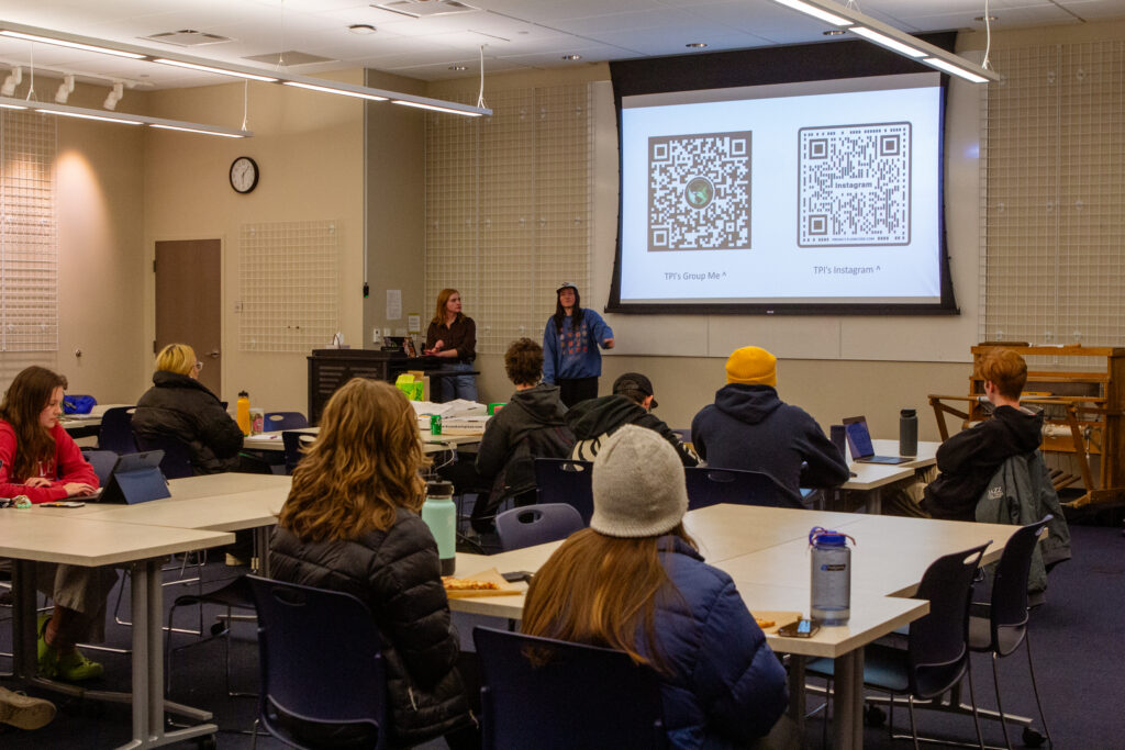 Students sit at tables and listen to someone speak.