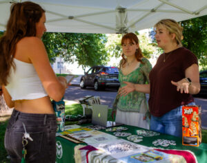 Two women speak to another woman outside underneath a tent.